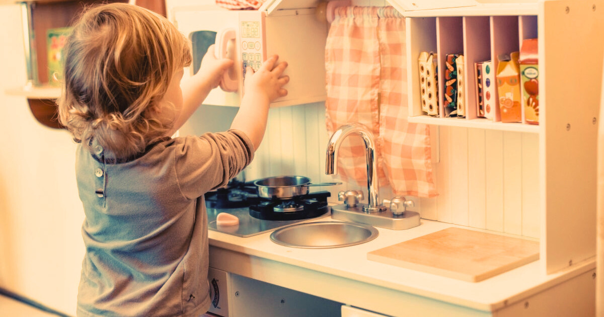 Toddler playing with wooden play kitchen putting dishes in cupboard